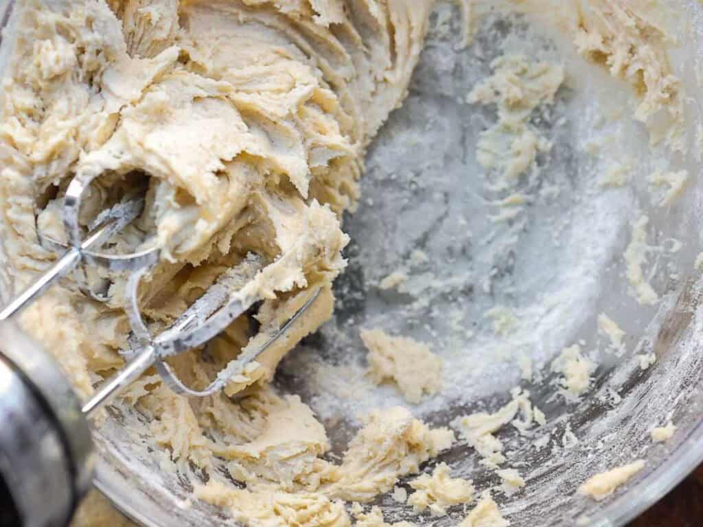 A close-up of electric mixer beaters blending creamy batter in a glass bowl, with some unmixed flour visible around the edges.